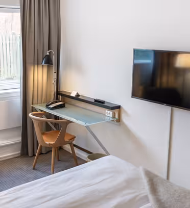 A modern hotel room with a neatly made bed, a wall-mounted TV, and a glass-topped desk. Theres a wooden chair and a lamp on the desk. A window with closed curtains is on the left, next to a blue armchair. The room has light-colored walls.
