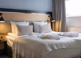 A cozy hotel room with a neatly made double bed, featuring plush white pillows and a soft cream-colored blanket. Two reading lamps are mounted on the wooden headboard, and robes are placed on the bed. A window with curtains provides natural light.