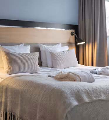 A cozy hotel room with a neatly made double bed, featuring plush white pillows and a soft cream-colored blanket. Two reading lamps are mounted on the wooden headboard, and robes are placed on the bed. A window with curtains provides natural light.
