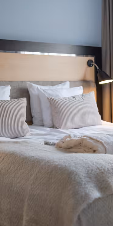 A cozy hotel room with a neatly made double bed, featuring plush white pillows and a soft cream-colored blanket. Two reading lamps are mounted on the wooden headboard, and robes are placed on the bed. A window with curtains provides natural light.