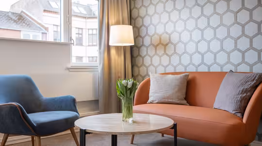A cozy living room with a blue chair, an orange sofa, and a round wooden coffee table. A vase of tulips sits on the table. A floor lamp is beside the sofa, and a patterned wallpaper covers the wall. A window shows a view of neighboring buildings.