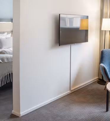 Modern hotel room with a view into a bedroom featuring a neatly made bed with white and gray bedding. The main area has a wall-mounted TV, blue armchair, round wooden table with a vase of tulips, and a standing lamp. Neutral decor and carpeted flooring.