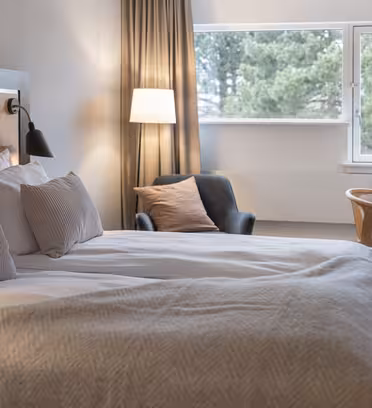 A cozy hotel room with a large bed, white pillows, and a beige throw blanket. Theres a gray lounge chair with a brown pillow, a wooden desk with a lamp, and a window with curtains letting in natural light. A vase and basket are on the desk.