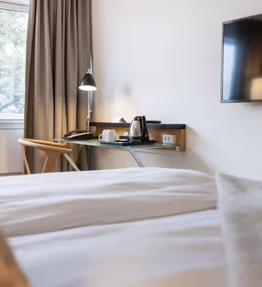 Modern hotel room with a neatly made bed, a wall-mounted flat-screen TV, and a window with closed curtains. A desk beside the window holds a lamp, kettle, cups, and a chair, creating a tidy workspace.