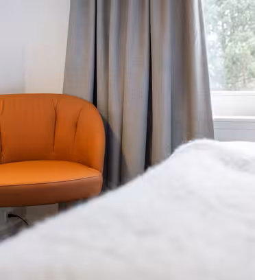 An orange armchair sits in a corner near a window with gray curtains. Soft daylight filters through the window, illuminating the room. A bed with a white blanket is partially visible in the foreground.