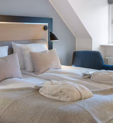 A cozy hotel room with a large double bed covered in a beige blanket and several pillows. Two robes are neatly placed on the bed. A modern lamp hangs above the bed, and a small window reveals part of a building outside. A blue chair sits nearby.