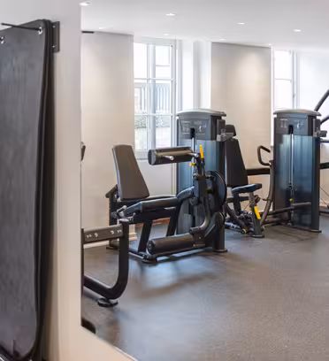 A modern gym with various exercise machines, including leg press and rowing machines. The room has bright lighting, light grey walls, and a dark, speckled floor. Large windows allow natural light to enter the space.