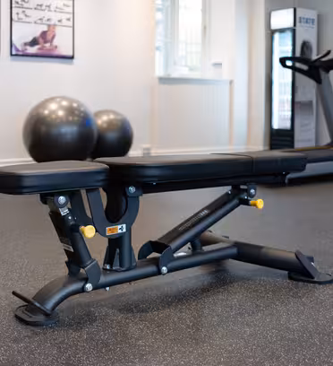 A modern gym with a black adjustable bench in the foreground. Behind it, two large exercise balls, a treadmill, and a rack with exercise equipment are visible. The gym has a light-colored floor and walls.