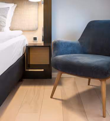 A modern bedroom features a blue upholstered mid-century chair with wooden legs, placed on light wooden flooring next to a neatly made bed with white bedding and beige pillows. A small, low wooden nightstand sits between the bed and the chair.