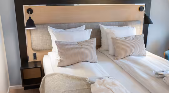 A cozy bedroom with two neatly made twin beds pushed together, each with white linens and striped pillows. The beds have a shared wooden headboard with two black reading lamps. A side table sits beside the beds under one lamp.
