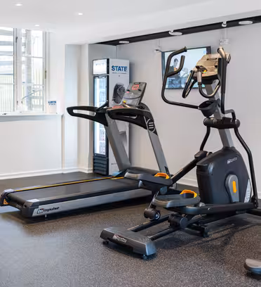 Small gym with a treadmill, elliptical machine, stationary bike, and exercise ball. A TV is mounted on the wall, and theres a window letting in natural light. A vending machine is also visible in the corner.