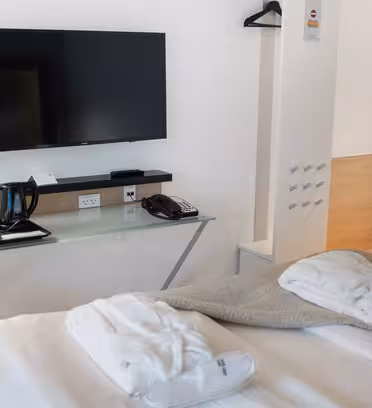 A modern hotel room with a neatly made bed topped with two white robes. A wall-mounted TV is above a glass desk holding a lamp, telephone, and tea-making essentials. A closet with hangers is on the wall, and the room has minimal, stylish decor.