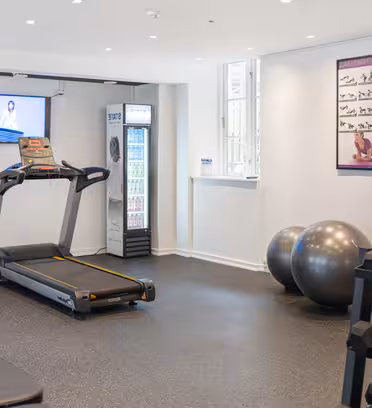 A small gym with a treadmill, an elliptical machine, two large exercise balls, and a rack of dumbbells. A large window lets in natural light. A TV is mounted on the wall, and there are exercise posters and a fridge with drinks.