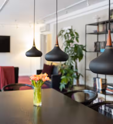A modern living room features three black pendant lights above a dining table with a vase of orange tulips. In the background are a TV, bookshelves, and a large plant. Dark chairs and maroon blankets accent the subdued décor.