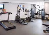 A modern gym with equipment including a treadmill, elliptical, stationary bike, and weight machines. The room has a rubber floor and white walls with a TV in the background.