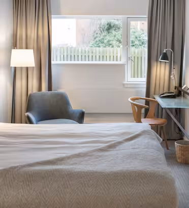 A modern hotel room features a neatly made bed with a light gray blanket, a cozy armchair, and a mounted flat-screen TV. A small desk with a phone and chair sits by a large window with gray curtains, allowing natural light to fill the space.