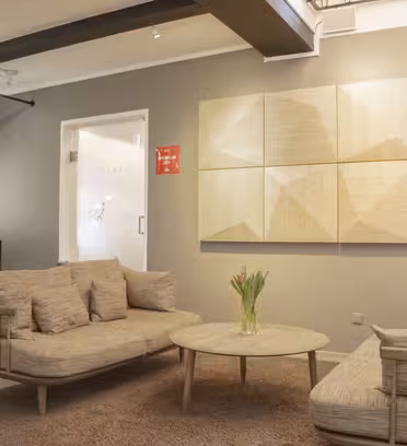 A cozy living room with a neutral color palette features two beige sofas and a round coffee table with a vase of tulips. Geometric wooden wall art hangs above, and a light rug covers the floor. A curtain and glass door are in the background.