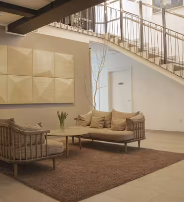 Cozy seating area with two beige sofas and a modern coffee table on a brown rug. Beige geometric wall art and potted plants add decor. An industrial-style staircase is in the background.