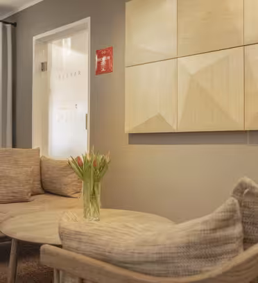 A cozy seating area with a beige sofa and armchair, a round wooden coffee table, and a vase of tulips. A geometric wooden art piece hangs on the wall next to a frosted glass door and long curtains.