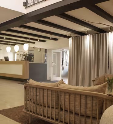 Spacious modern lobby with beige sofas and a large vase on a brown rug. Overhead beams and pendant lights illuminate the space. A reception desk with computers is in the background, and a spiral staircase is on the left. Neutral tones dominate the decor.