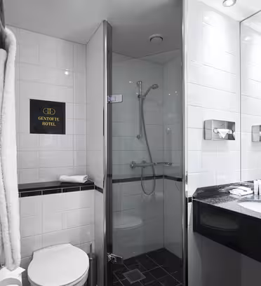 Modern hotel bathroom with white tiled walls, a glass-enclosed shower, a black granite countertop, and a round sink. Towels hang on the wall beside the toilet. Minimalist lighting and amenities are placed neatly on the sink.