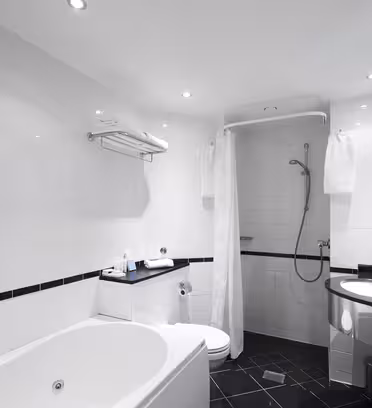 Modern bathroom with white tiles, featuring a bathtub, a wall-mounted shower with curtain, and a sleek sink on a granite countertop. Towels are neatly placed on a rack and shelf. Bright lighting and black floor tiles complete the clean look.
