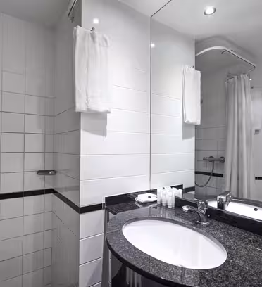 A modern bathroom with white tiled walls, a wall-mounted shower, and a black granite countertop with a sink. A large mirror reflects the space. Towels hang on a rail, and toiletries are neatly arranged by the sink.