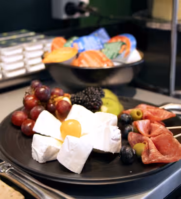 A black plate with a selection of cheese, salami, grapes, blackberries, blueberries, olives, and a cherry tomato. In the background, there are small cream containers and packets of butter.
