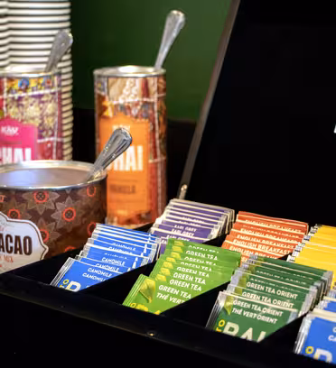 A variety of tea bags are organized in a black box labeled Lipton on a counter. Nearby are stacked paper cups, canned chai mixes, a bowl with tea packets, and a cocoa canister. The setup appears to be a beverage station.