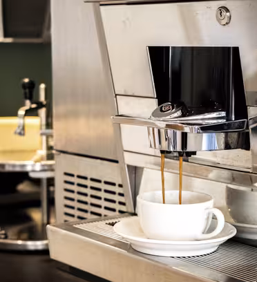 A modern espresso machine dispensing coffee into a white cup and saucer. The machine is silver, featuring a sleek design, with dark coffee flowing from the spout. A blurred background includes another beverage machine and shelves.