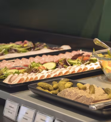 A buffet table with trays of assorted cold cuts, sliced cucumbers, cherry tomatoes, mixed greens, pickles, and a bowl of mixed fruit. Tongs are placed beside the dishes.