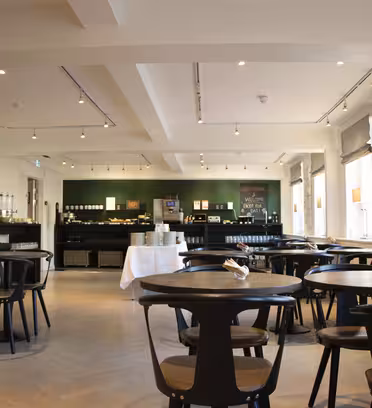 A modern, well-lit cafeteria with sleek black tables and chairs. There is a serving area in the back with food and beverages. Large windows on the right let in natural light, creating a welcoming atmosphere.