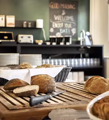 Bakery setting with fresh bread and pastries on wooden boards. A knife rests on sliced bread. Plates and glasses are neatly stacked on shelves in the background, beside a chalkboard with Good Morning & Welcome Enjoy Your Day! written on it.