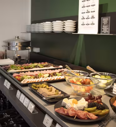 A buffet table with various dishes including sliced meats, cheeses, fruits, vegetables, and a stack of plates. There is a chafing dish and bowls with greens and tomatoes. The decor includes a green and black color scheme with a small sign on the wall.
