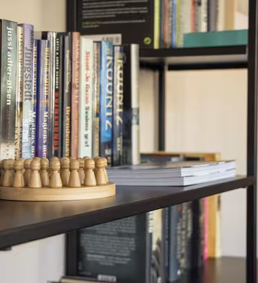 A bookshelf filled with various books and magazines. In the foreground is a wooden chess piece set with pawns on a round board. Some books are stacked vertically, while others are laid flat. The shelves are black metal.