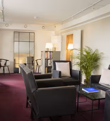 A modern lounge with black leather chairs and a sofa surrounding a dark coffee table. A tall plant and bookshelves adorn the room. Abstract art hangs on the walls, and purple carpeting complements striped curtains covering the windows.